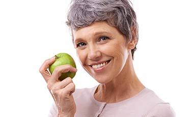 Smiling woman with dental implant supported replacement teeth holding a green apple
