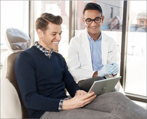 Man in sweater and dentist in lab coat smiling, seated in a bright dental office, looking at a tablet together, conveying friendliness and comfort.