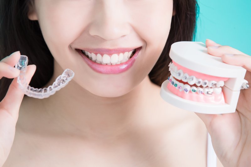 a woman smiling while holding braces and Invisalign aligners